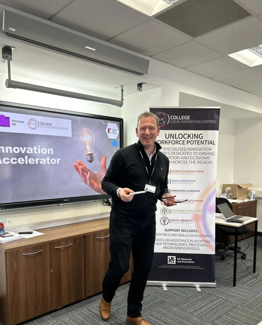 Presenter smiling at the College Local Innovation Centres event at Glasgow Kelvin College, standing in front of a banner and innovation display. Presenter smiling at the College Local Innovation Centres event at Glasgow Kelvin College, standing in front of a banner and innovation display.